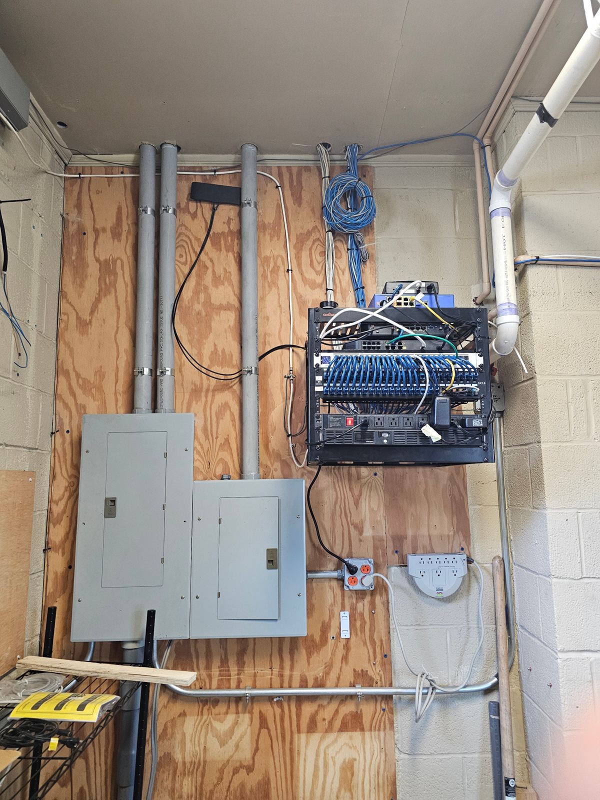 Wall-mounted network rack with a patch panel of blue Cat6 cables, switch and PDU, installed next to electrical panels